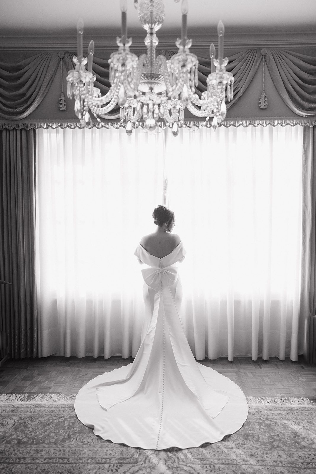 Silhouette of a bride in white gown standing beneath crystal chandelier with elegant draped curtains in black and white.