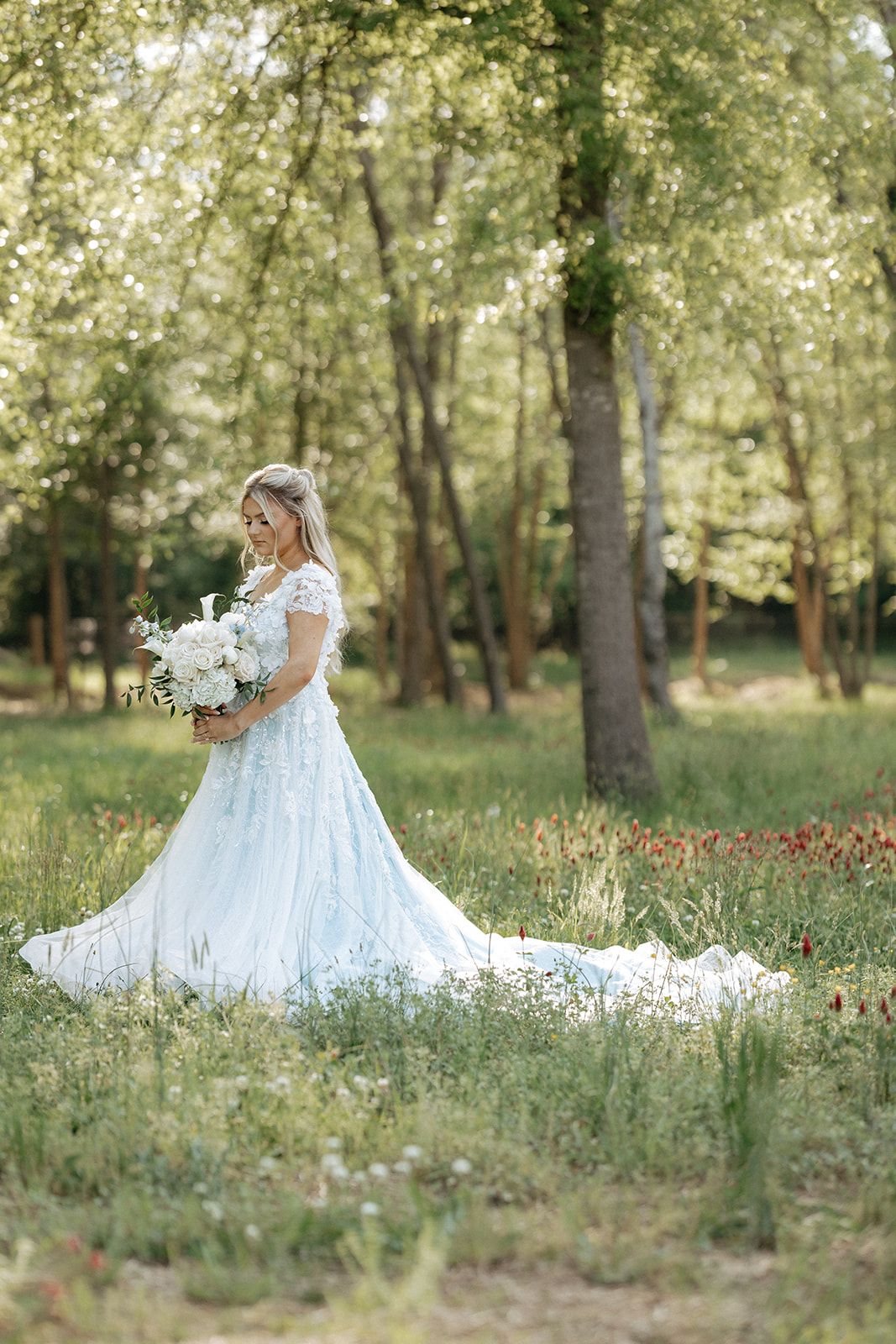A bride in a flowing white lace wedding gown stands gracefully in a sunlit forest holding a white floral bouquet.