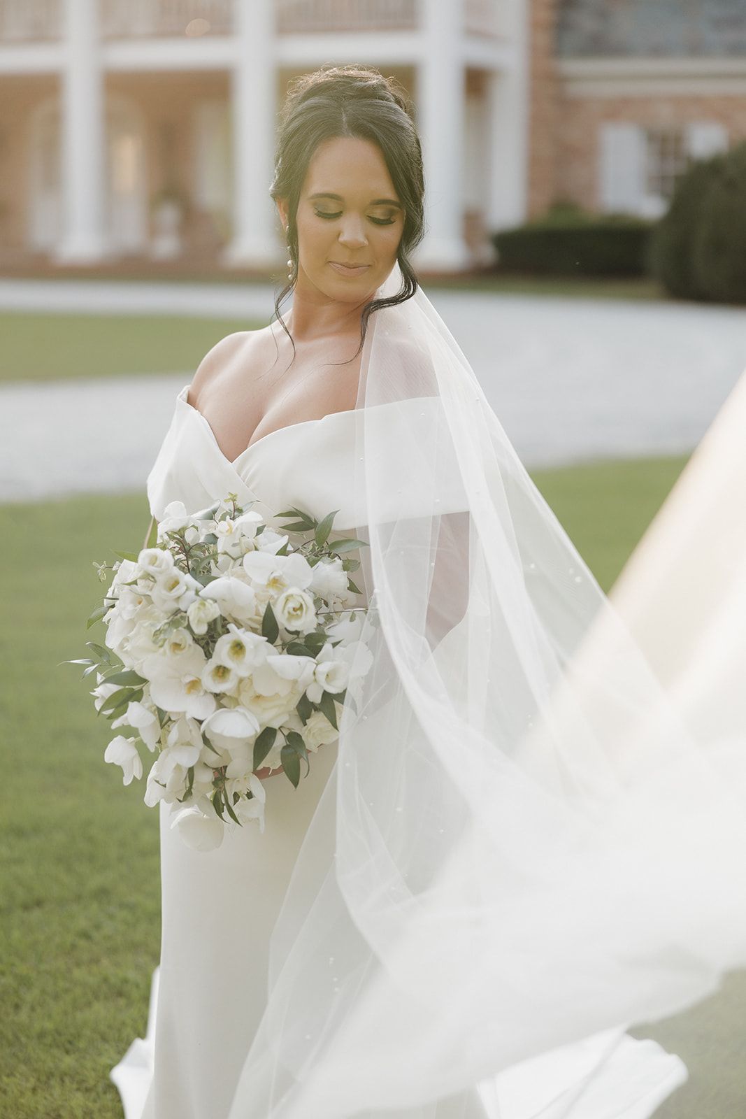 A beautiful bride in a sleek white gown holds an elegant bouquet of white flowers as her veil flows in the breeze.