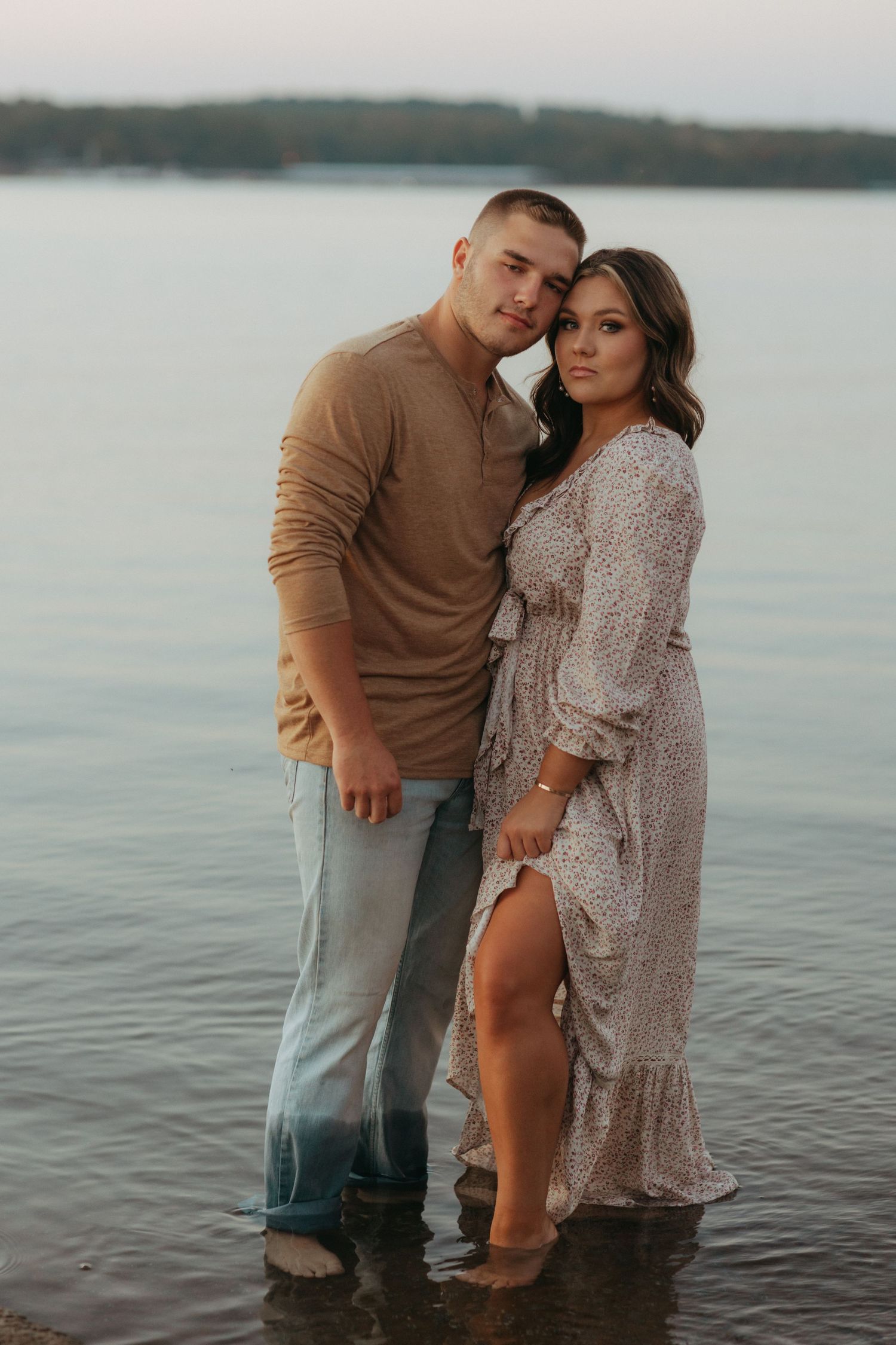 Couple stands in water at sunset, embracing in coordinated brown and beige outfits.