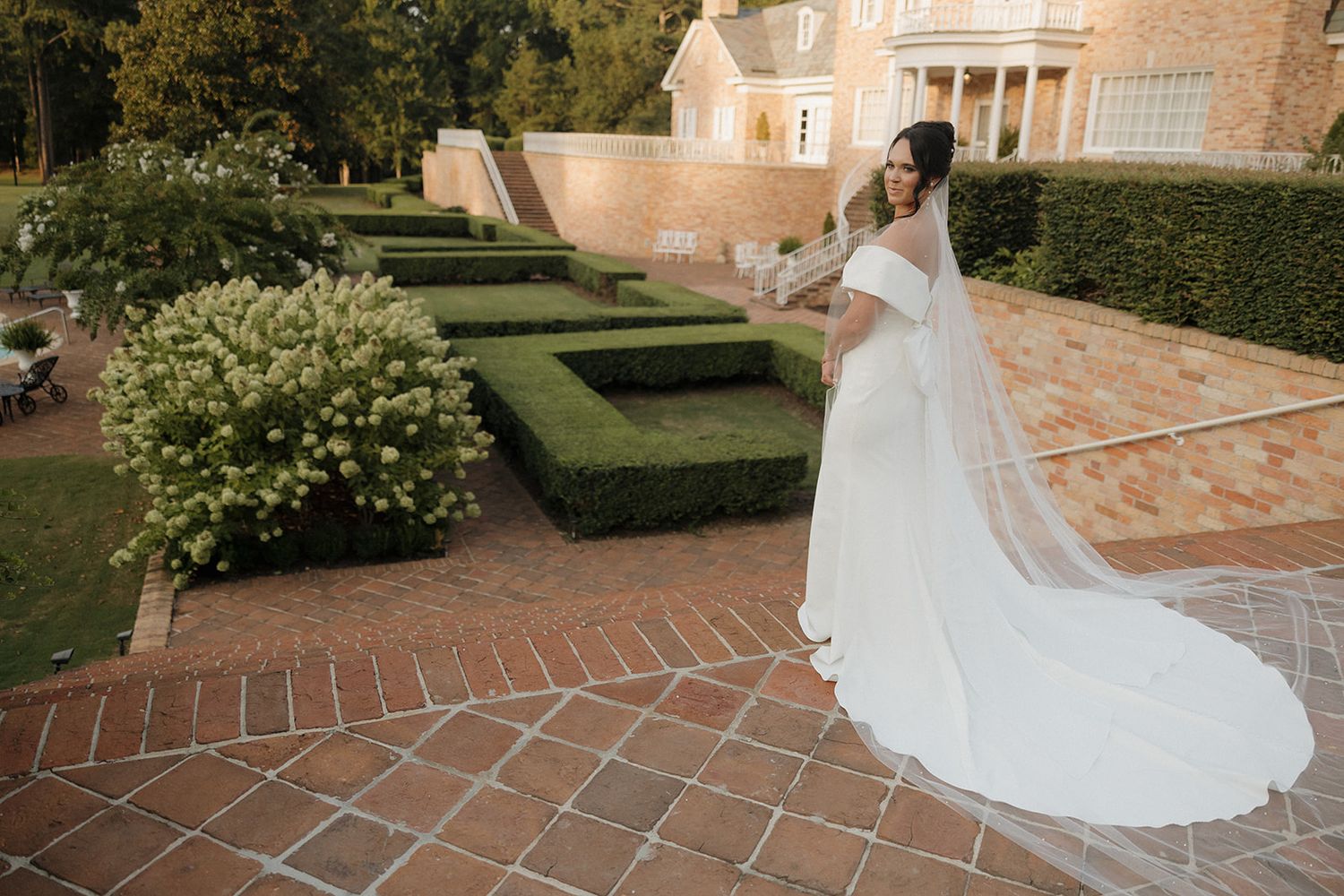 A flowing white wedding gown trails elegantly across sun-drenched bricks at a grand estate venue.