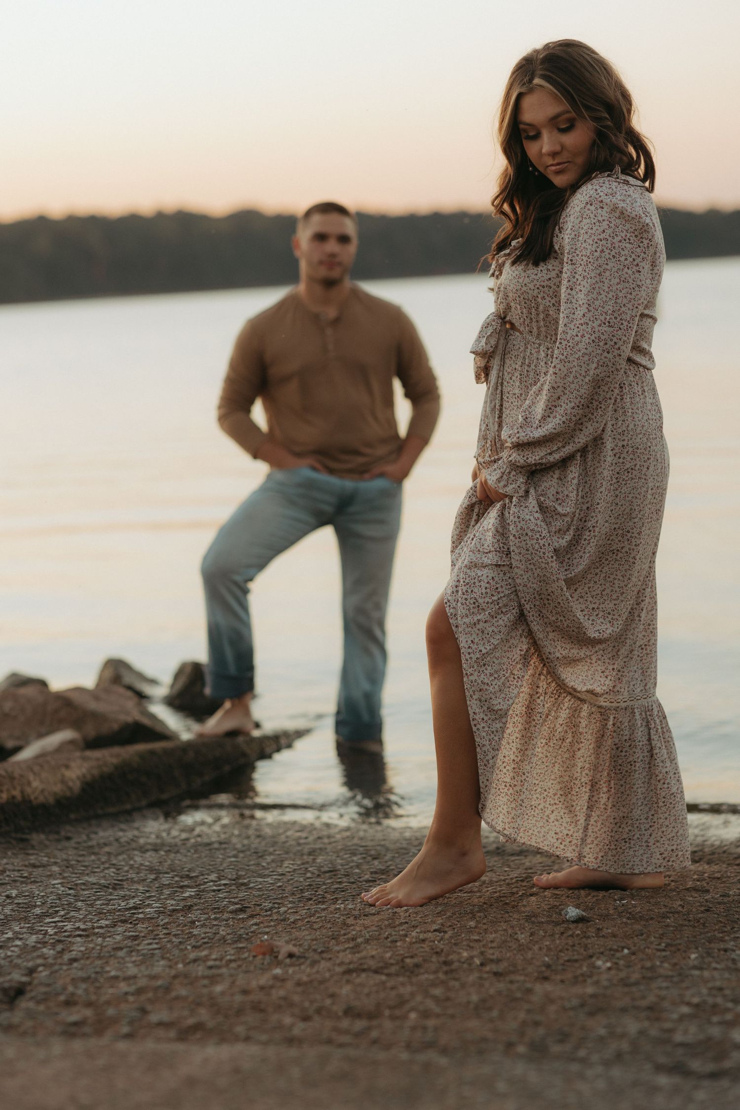 Couple poses by lakeside at sunset, woman in lace dress with man standing in background.