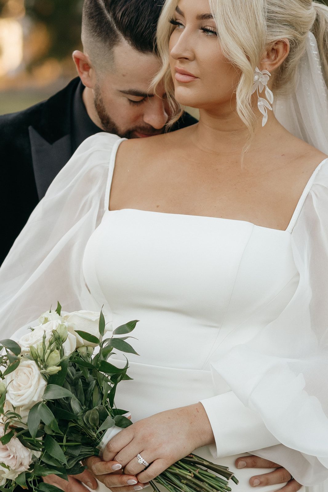 Bride in modern white dress with long sleeves holds a bouquet of white roses and greenery.