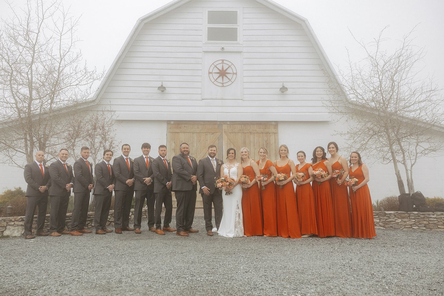 Wedding party members in alternating black and orange formal attire pose in front of a white barn on a misty day.