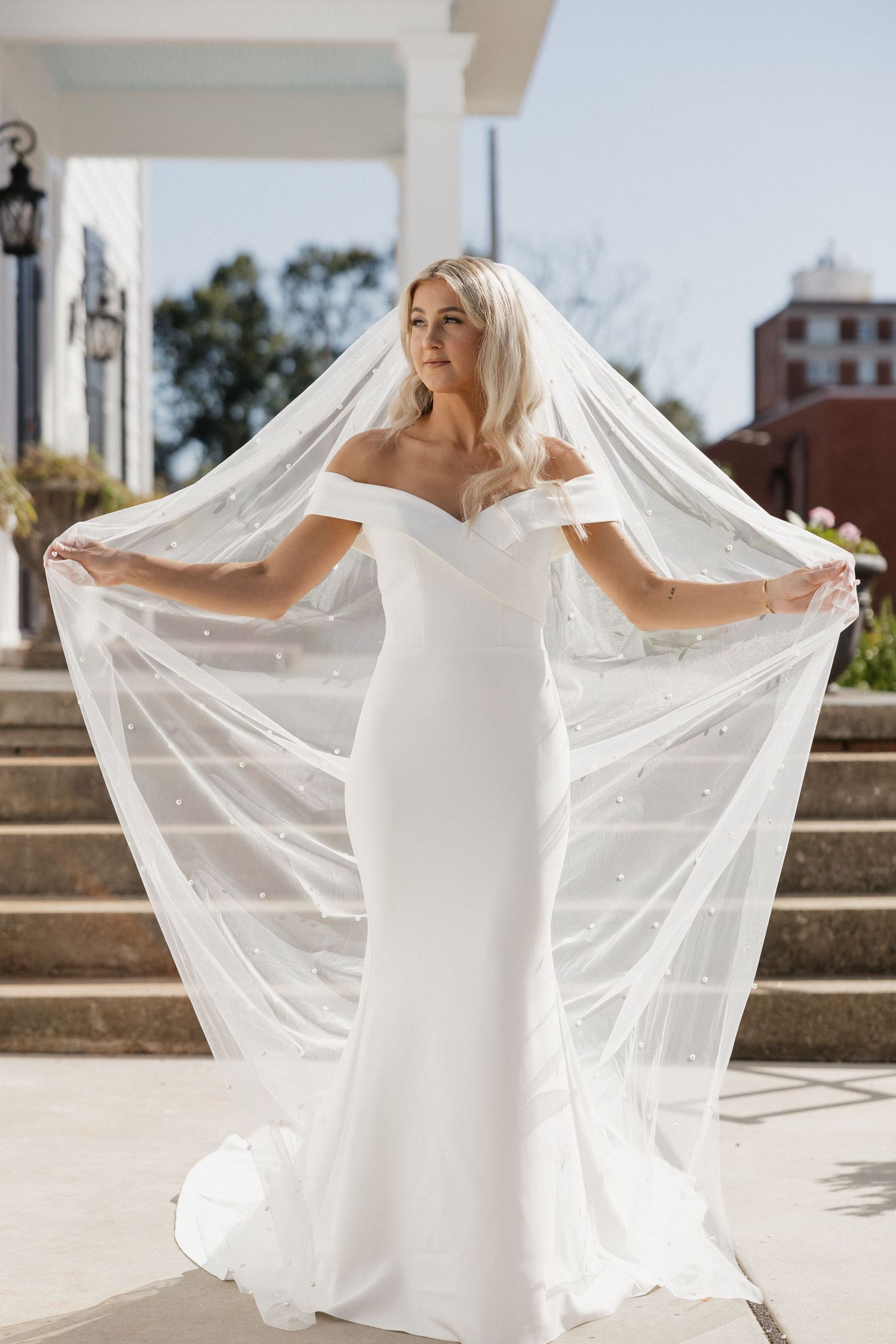 Someone wearing a flowing white wedding dress stands on stone steps as sunlight illuminates their long sheer veil.