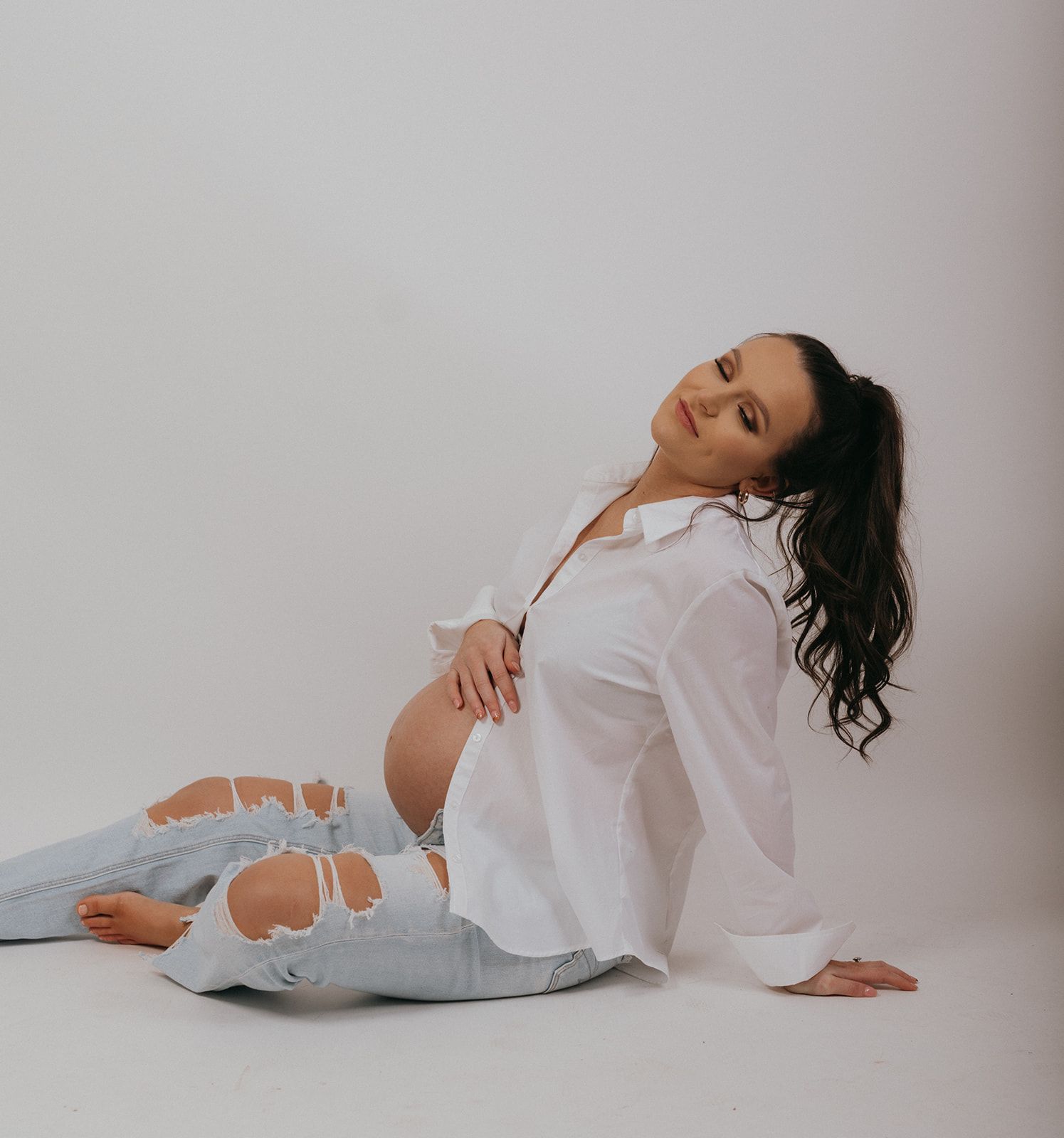 A pregnant woman in distressed jeans and white button-up shirt poses elegantly against a light studio background.