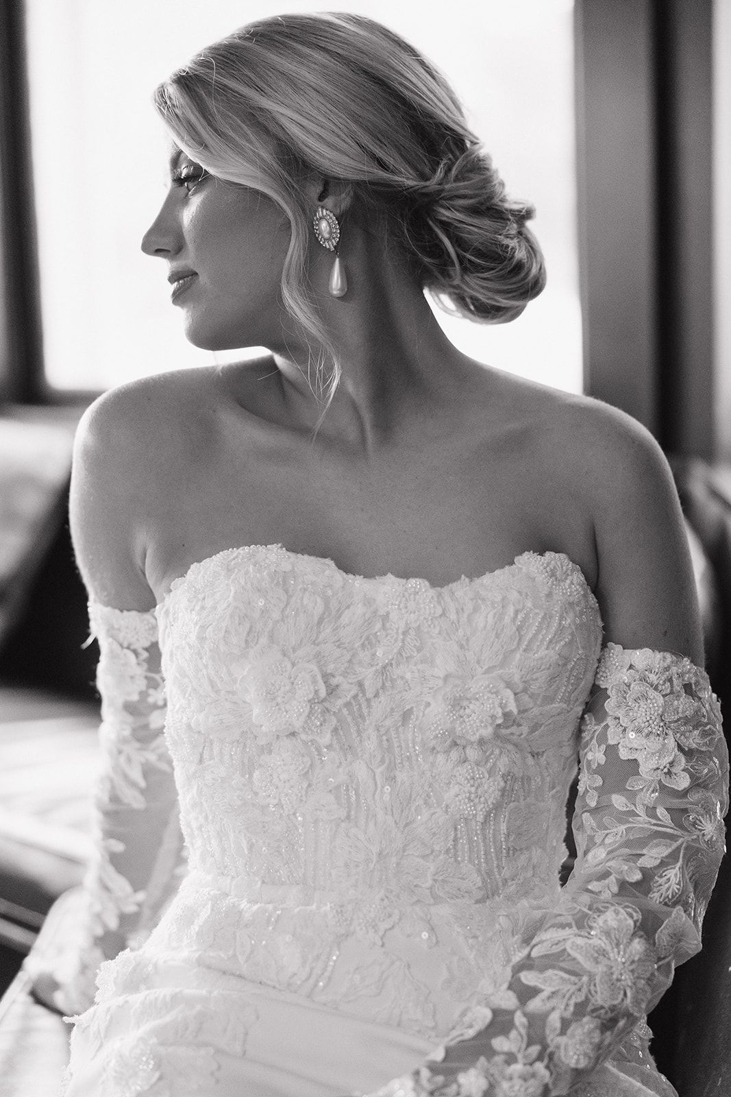 Black and white portrait of a bride in a lace off-shoulder wedding gown with delicate long sleeves looking to the side.