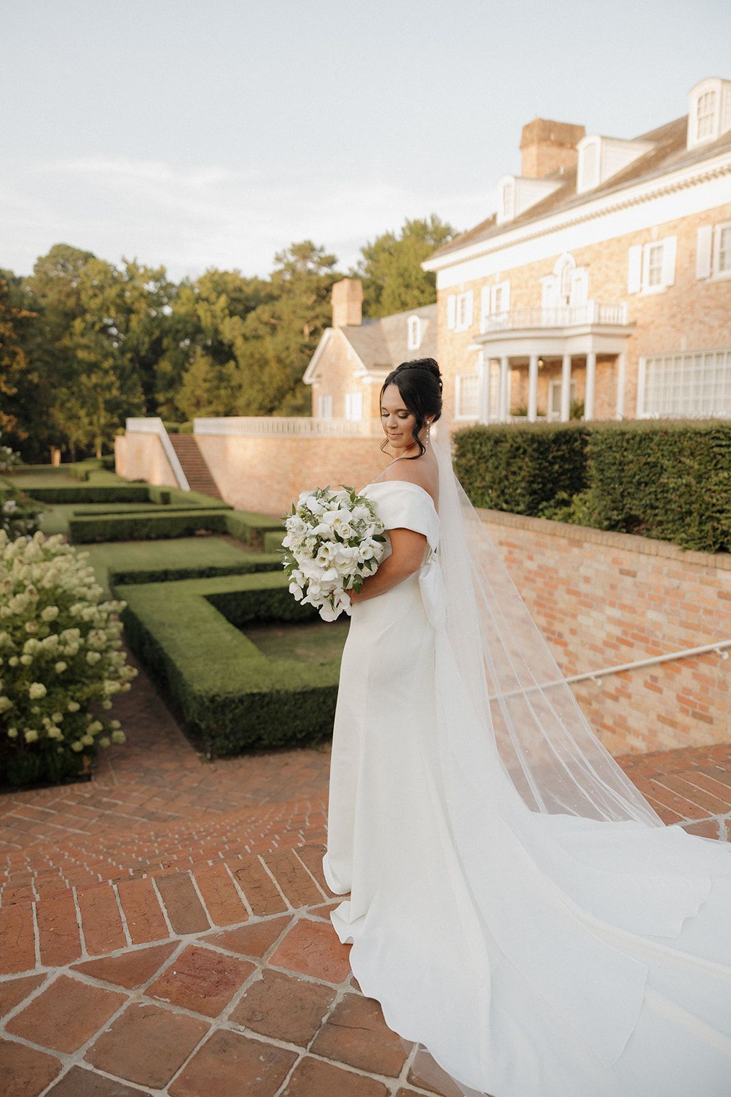 Elegant bride in off-shoulder gown holding white bouquet poses in formal garden with classic mansion in background.