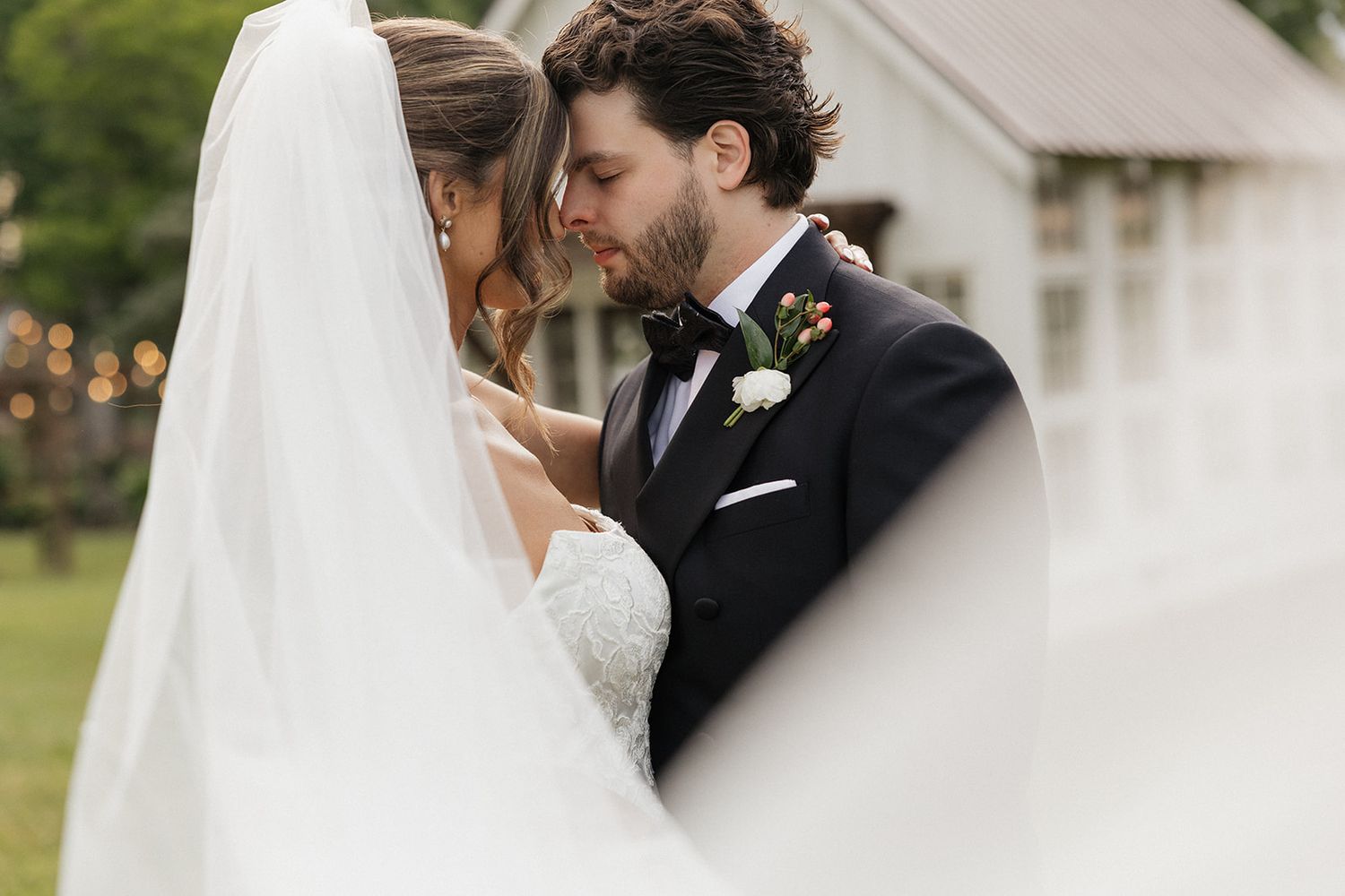 A newlywed couple shares an intimate moment outdoors as the bride's veil flows in the foreground.