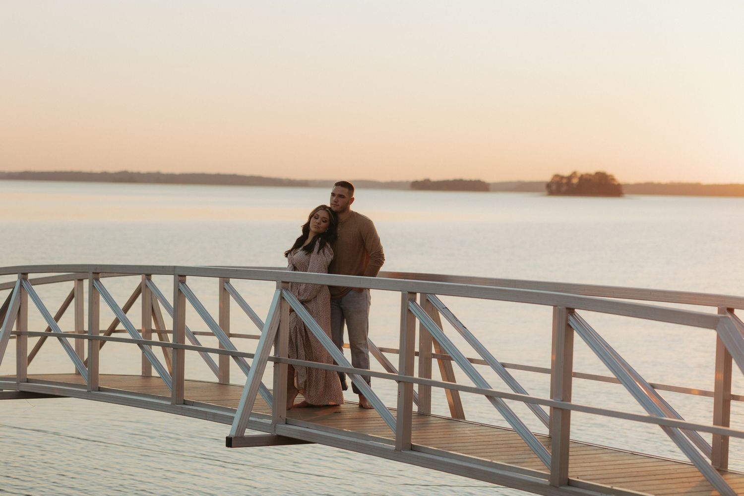 Silhouetted couple stands together on pier during golden hour overlooking calm lake.