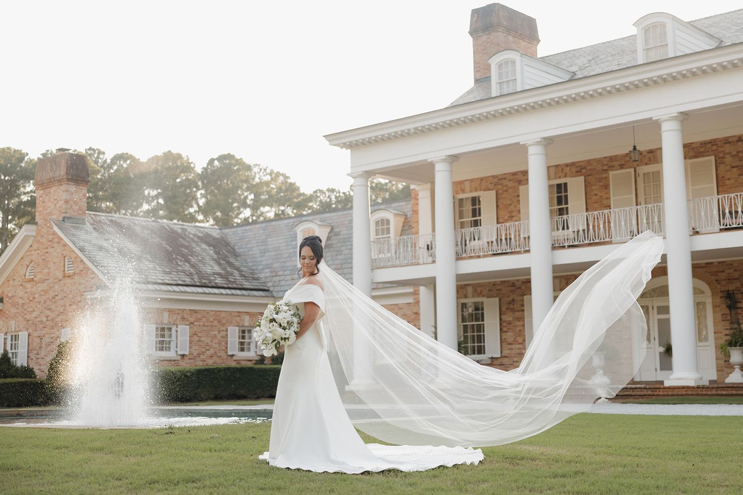 A bride in a flowing white wedding dress with a dramatic long veil stands on a lawn in front of a grand Southern mansion.