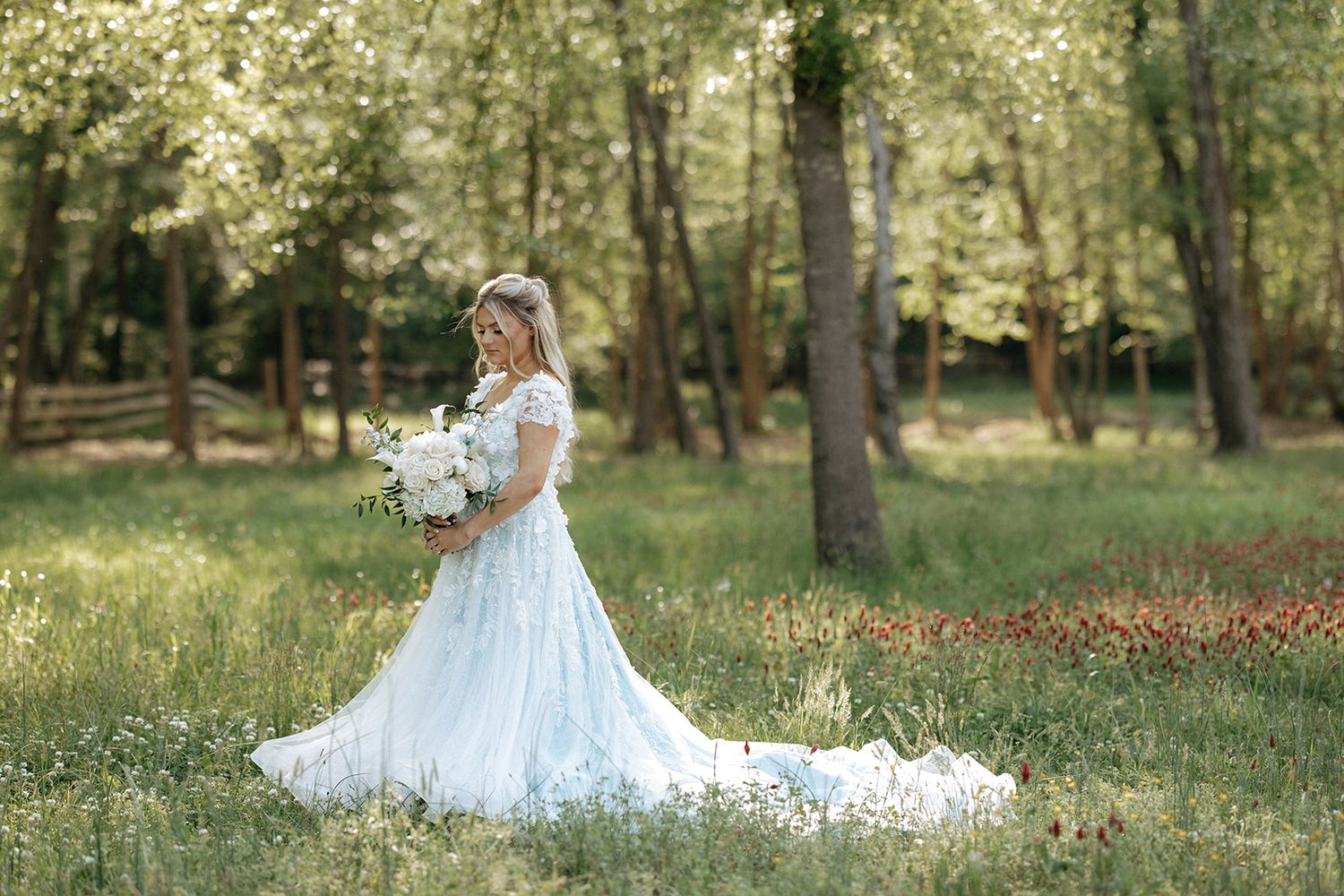 A bride in a flowing white lace gown holds a bouquet while standing in a sun-dappled forest clearing.