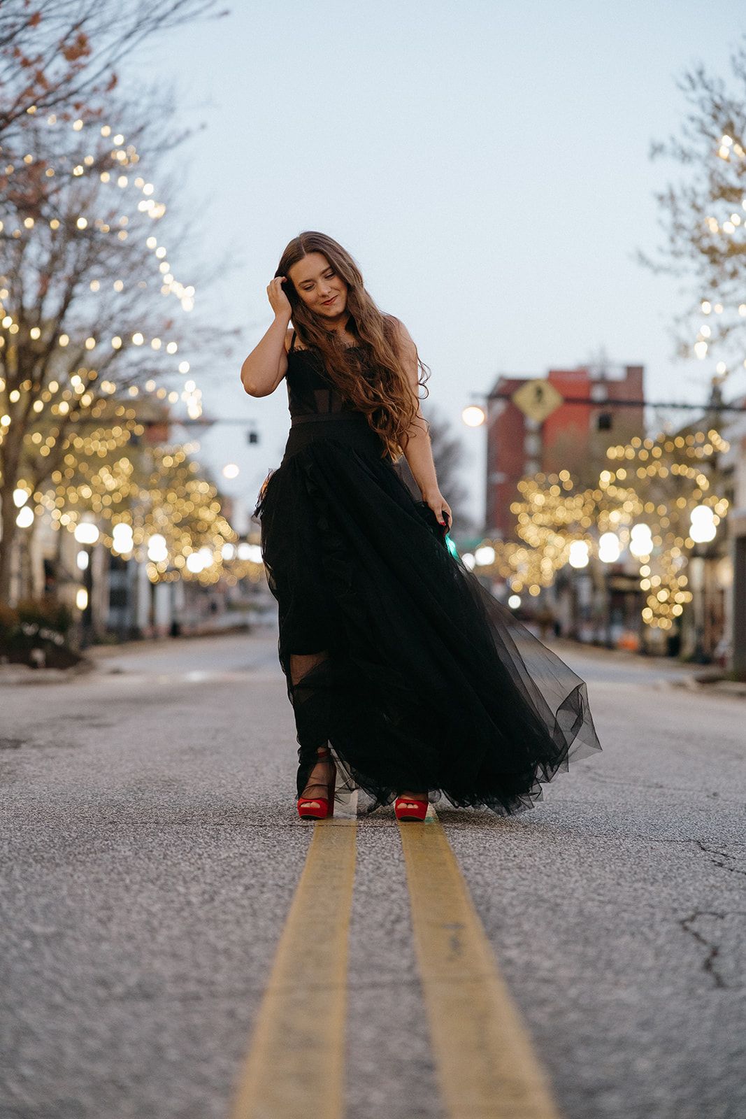 Figure in flowing black tulle gown stands on double yellow road lines at dusk with decorative street lights glowing.