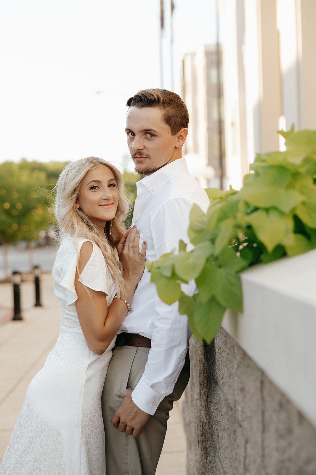 A romantic wedding couple poses together against a light-colored outdoor colonnade with greenery.