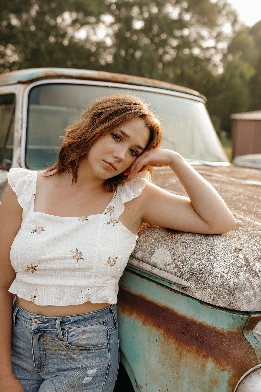 A person in a white crop top and denim jeans poses against a vintage rusty turquoise truck in warm sunset lighting.