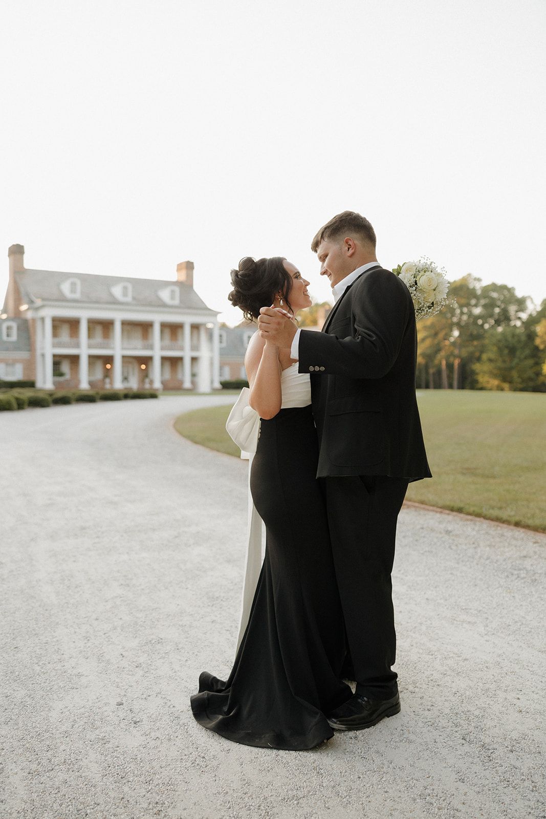 A couple shares an intimate moment in formal wear in front of a stately mansion.