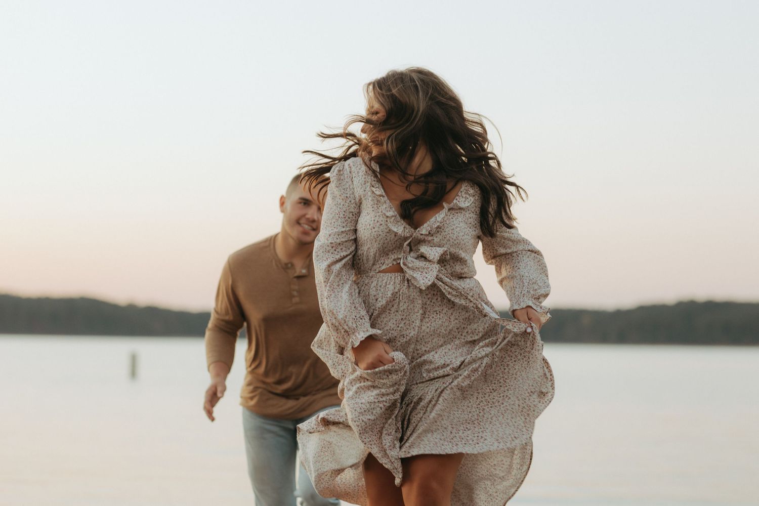 Romantic couple sharing a joyful moment by the water with wind-swept dress and hair.