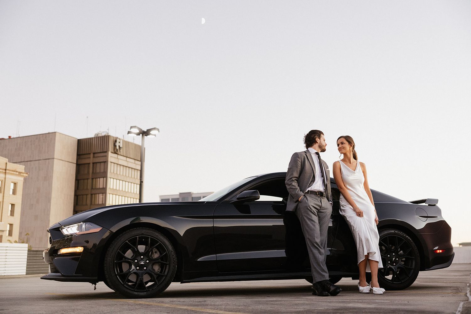 Stylish couple poses next to black sports car on urban rooftop.