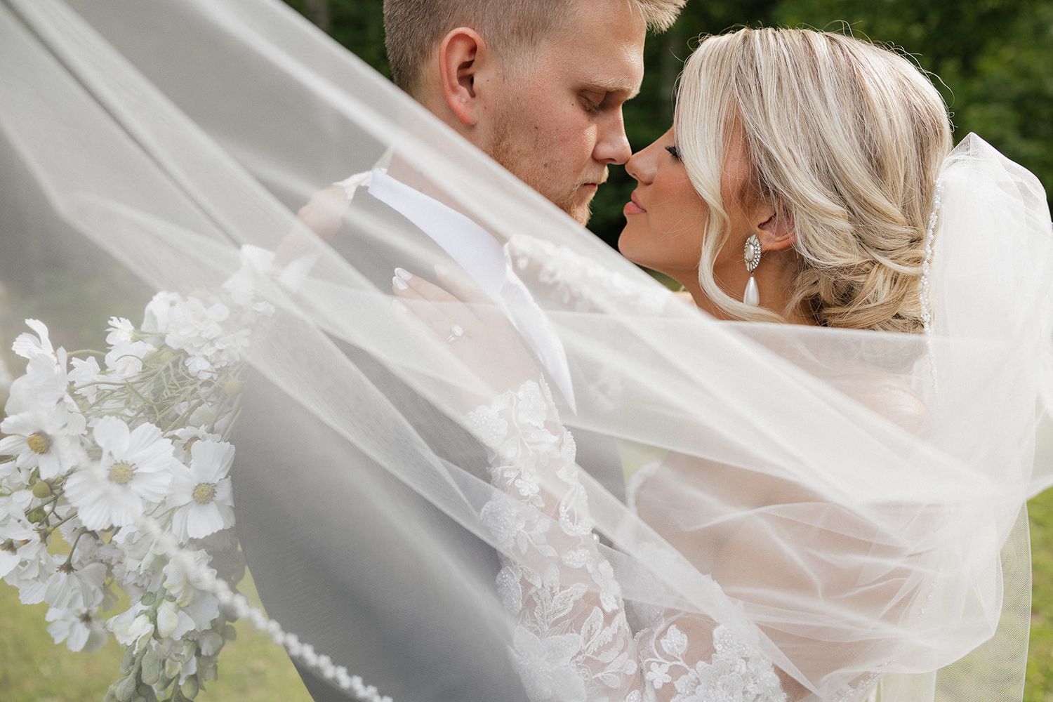 Romantic close-up of newlywed couple embracing while bride holds white floral bouquet under flowing veil.