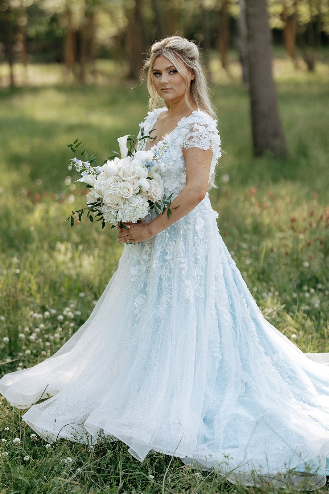 Bride wearing ethereal light blue tulle wedding gown with lace details holds white floral bouquet in sunny garden setting.