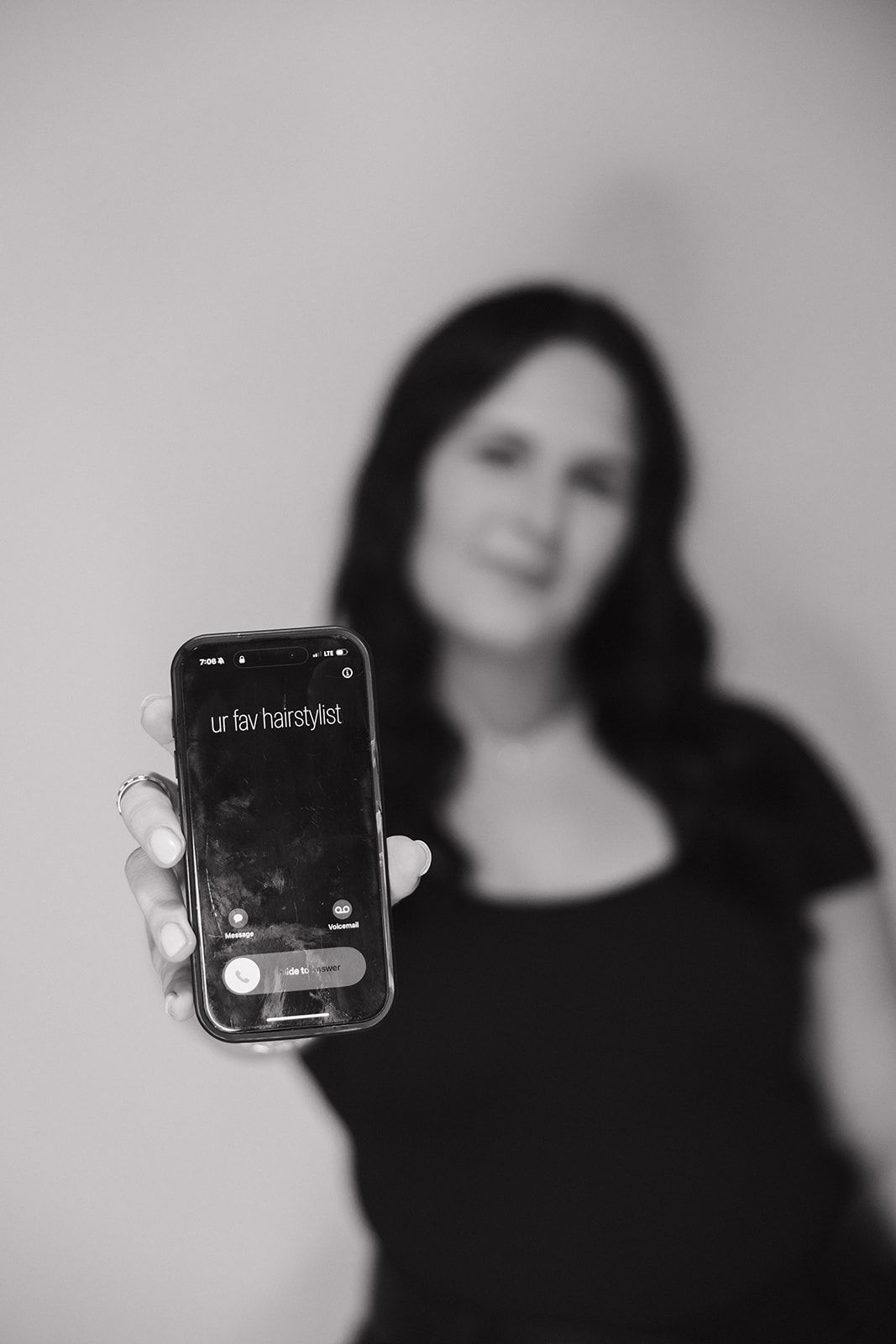 Black and white portrait of someone holding an iPhone displaying 'ur hairstylist' text on screen against blurred background.