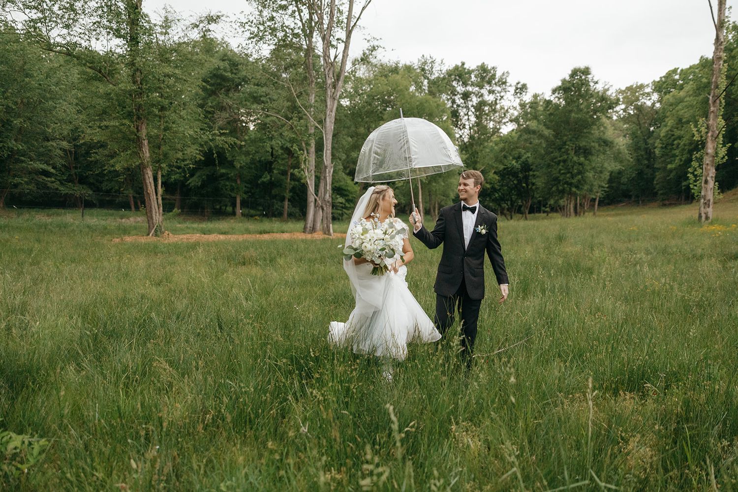 Newlyweds walk together through rainy field holding clear umbrella.
