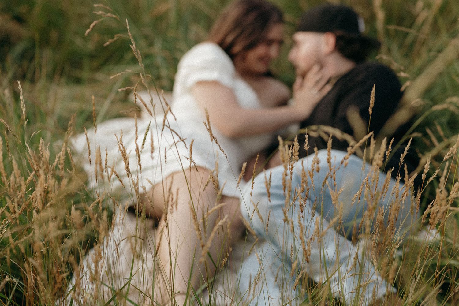 A romantic couple shares an intimate moment while lying in tall golden grass during a countryside photoshoot.