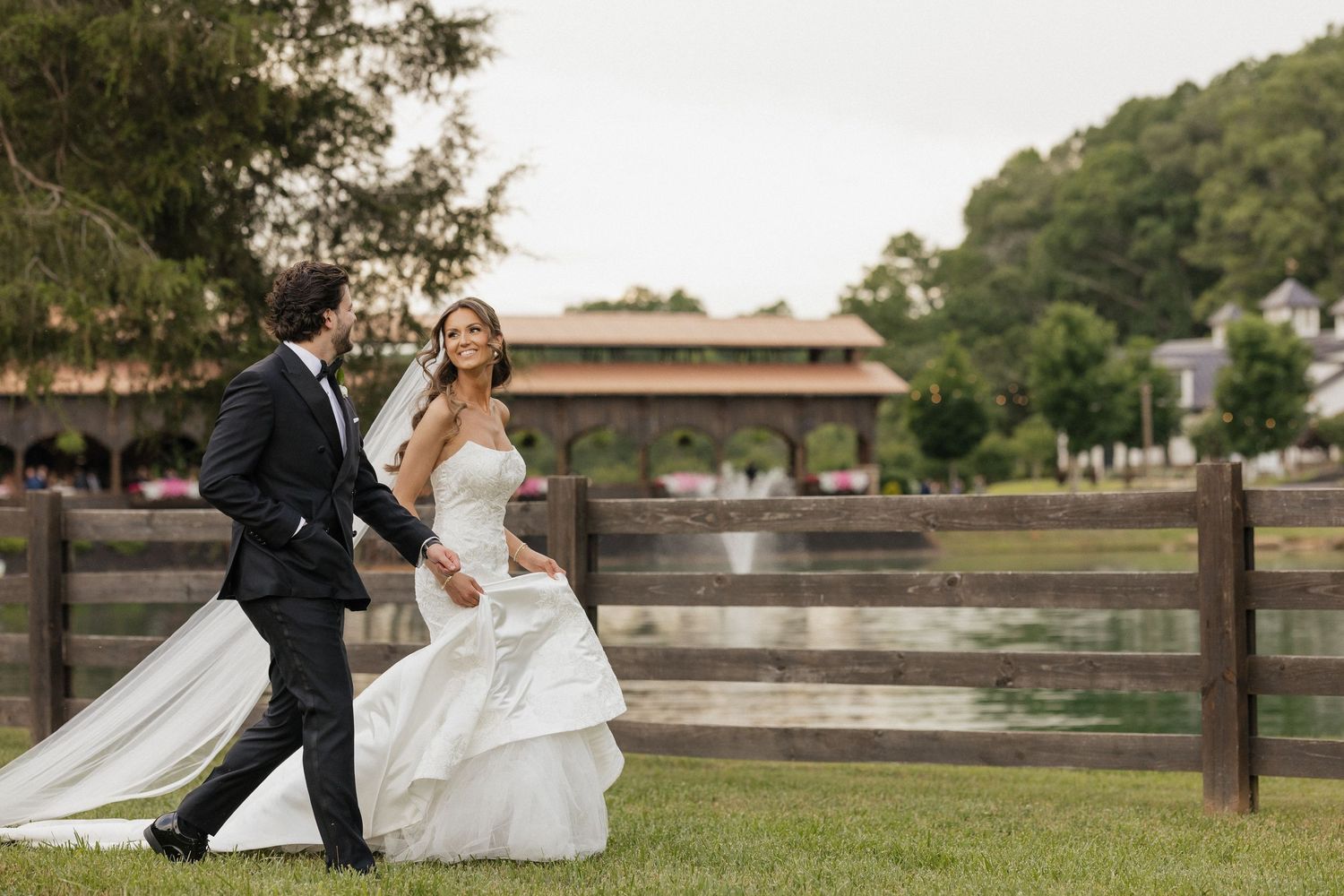 A couple in wedding attire walk together on a grassy lawn alongside a wooden fence at an outdoor rustic venue.
