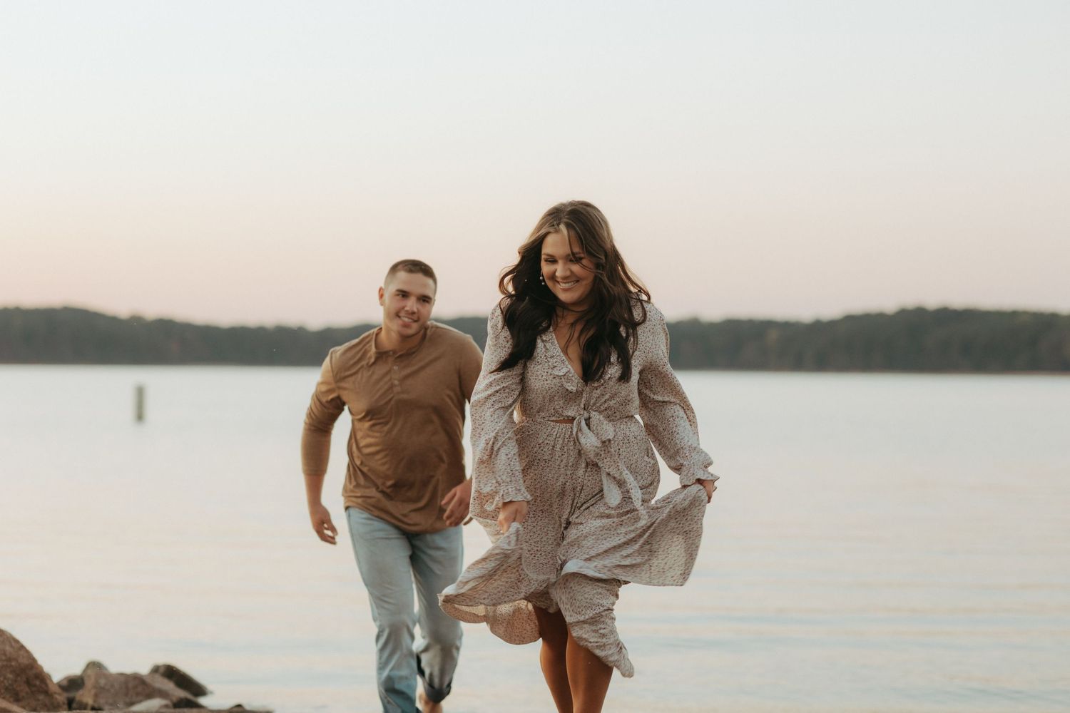 Playful couple running along rocky shoreline at sunset in casual romantic photoshoot.