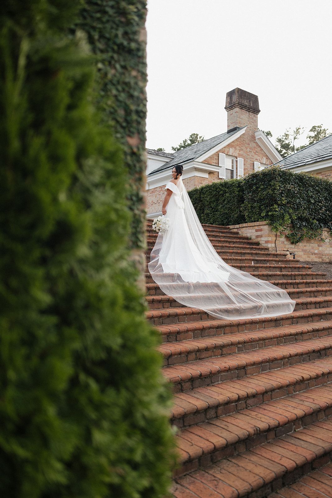 Flowing white wedding gown and veil cascade down elegant brick stairs at a historic mansion with ivy-covered walls.
