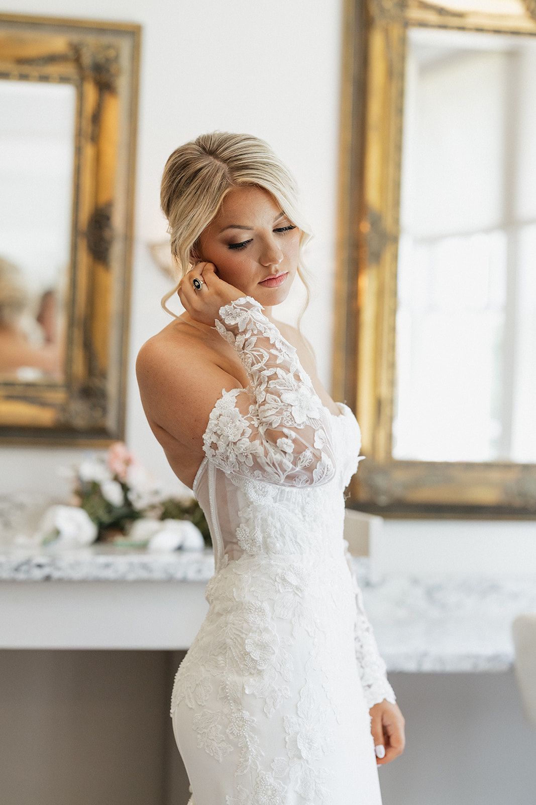 Bride in lace-sleeved wedding dress poses elegantly beside gold-framed mirror.
