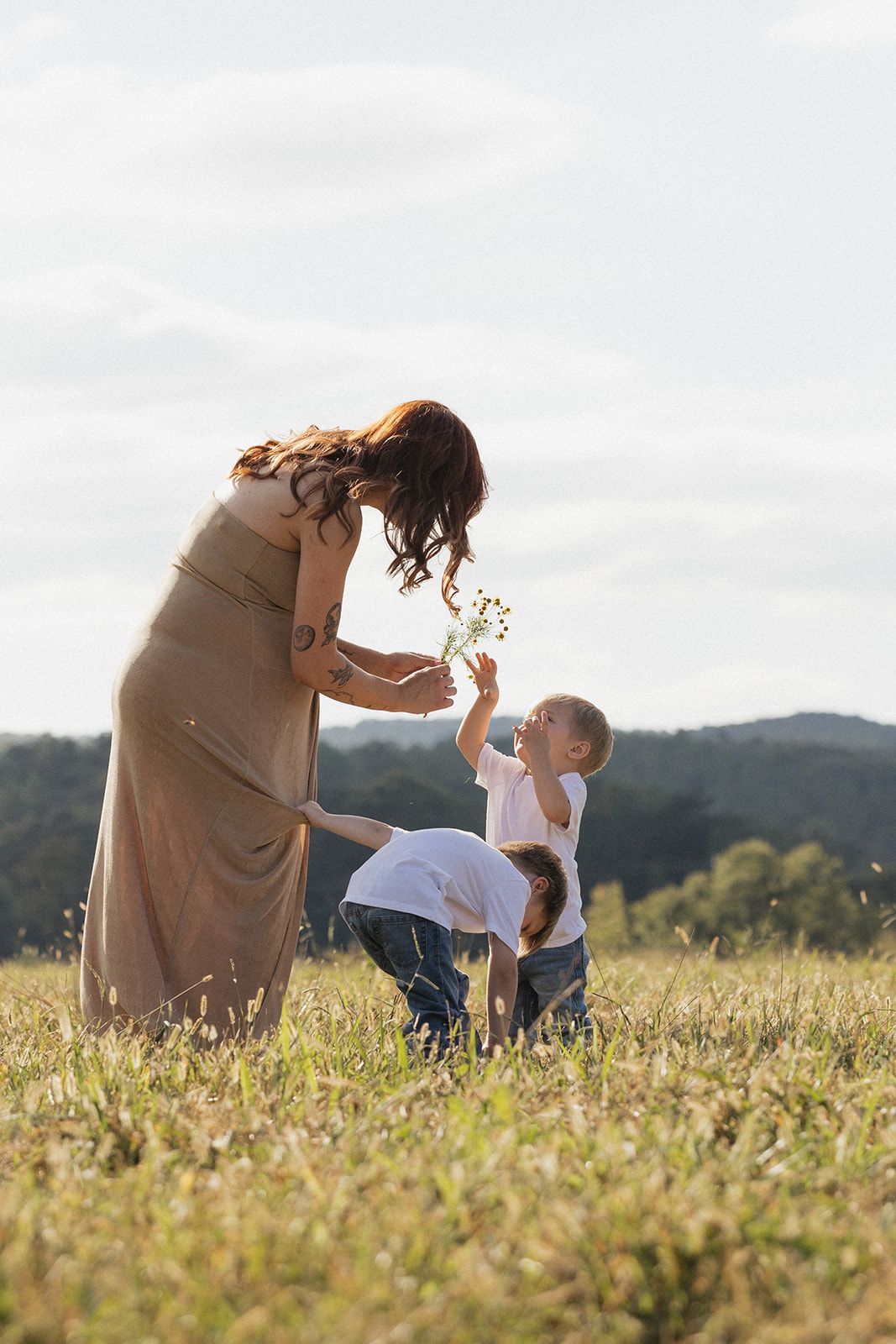 A woman in a flowing brown dress shares a tender moment with a young child in a sunlit meadow against rolling hills.
