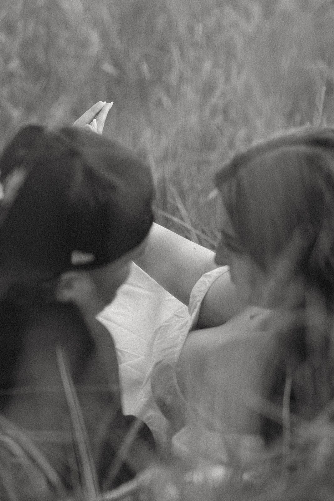 Two figures sit close together reading a book in tall grass, captured in black and white.