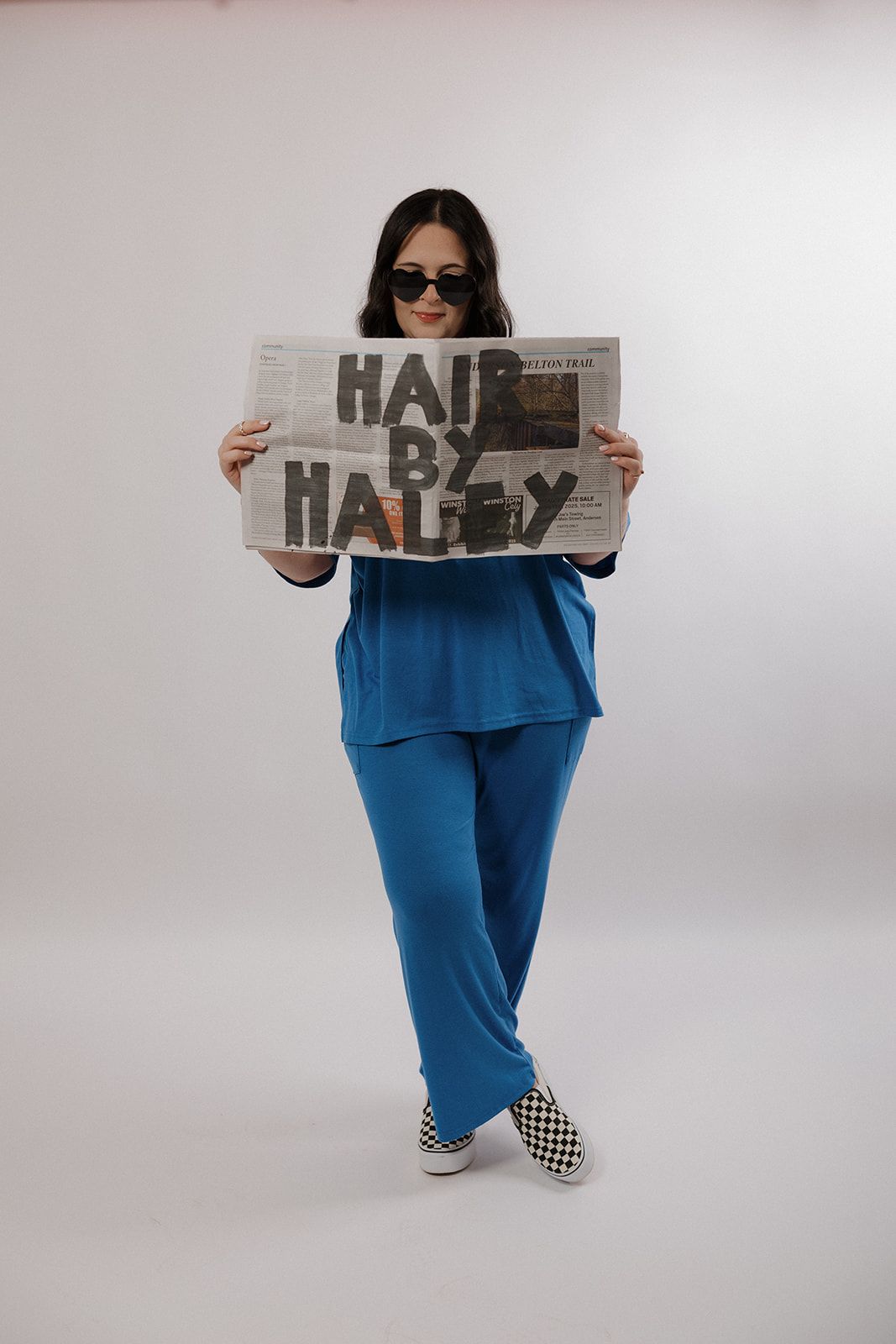 A person in a bright blue outfit and sunglasses holds up a newspaper while standing against a white backdrop.