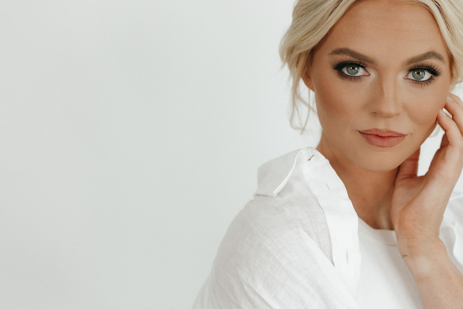 Person wearing a white button-down shirt poses elegantly against light background with soft makeup and styled blonde hair.