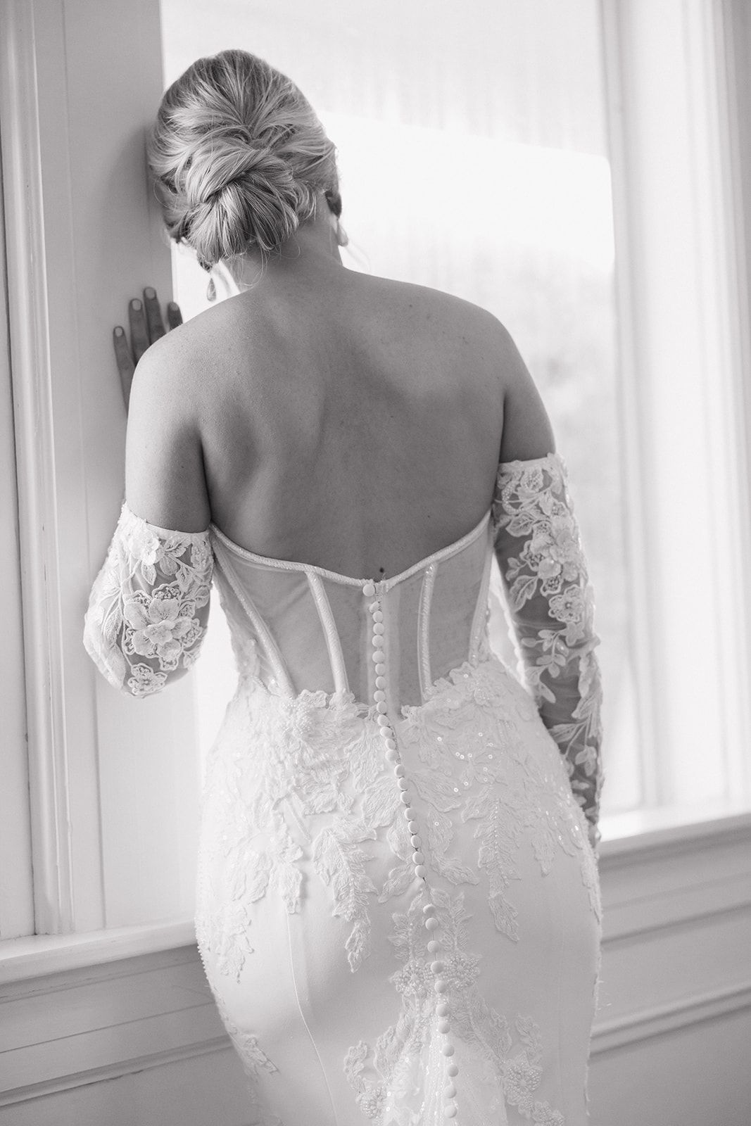 Back view of a bride in a fitted lace wedding dress with corset top and delicate button details, standing by a window.