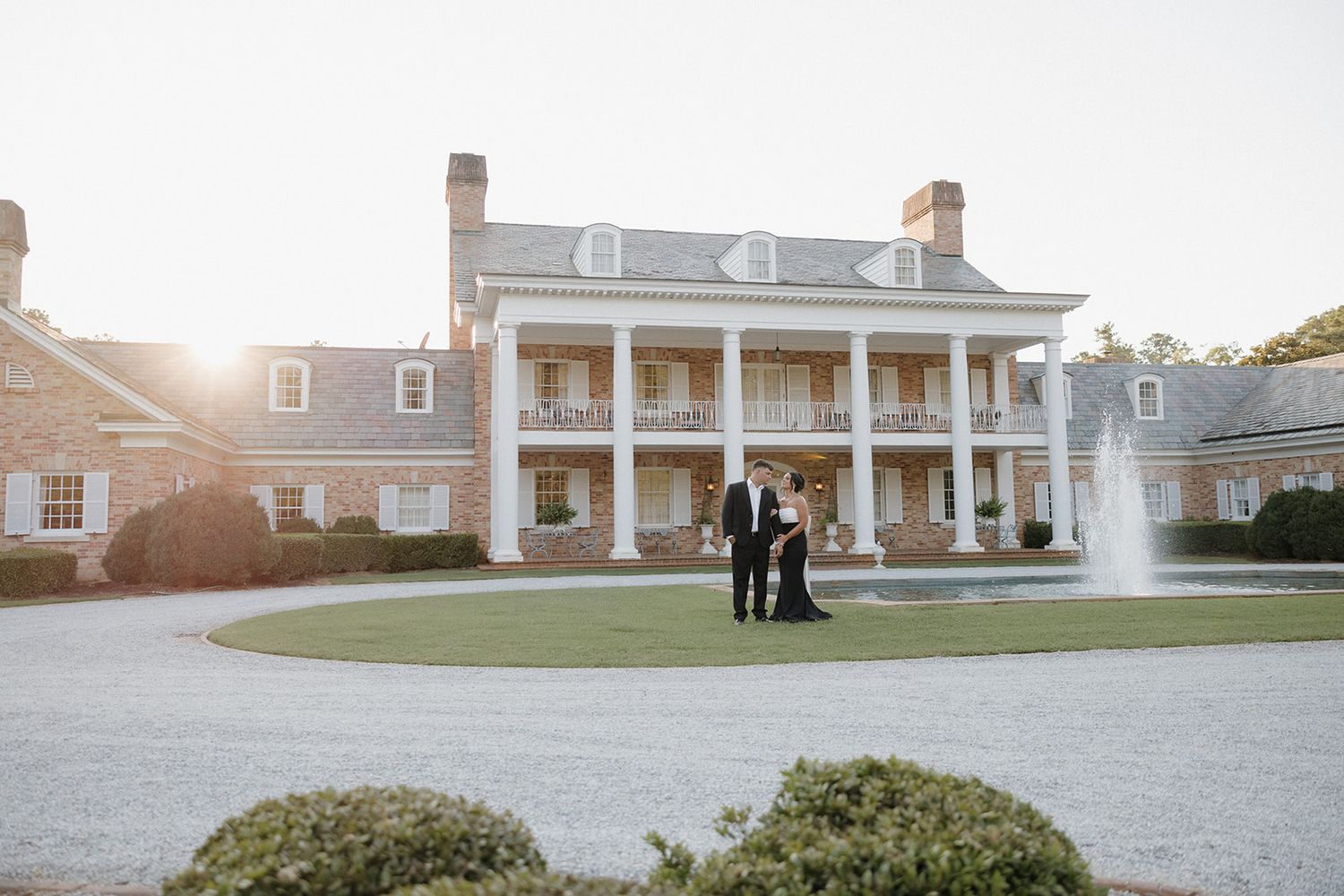 Wide panoramic view of a stately brick mansion with white columns and fountain.