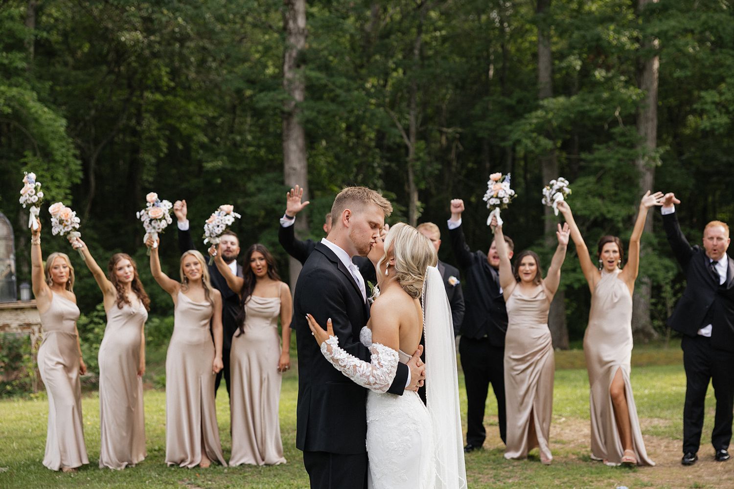 Wedding party in formal attire celebrates outdoors, standing in a line while raising their hands joyfully in unison.