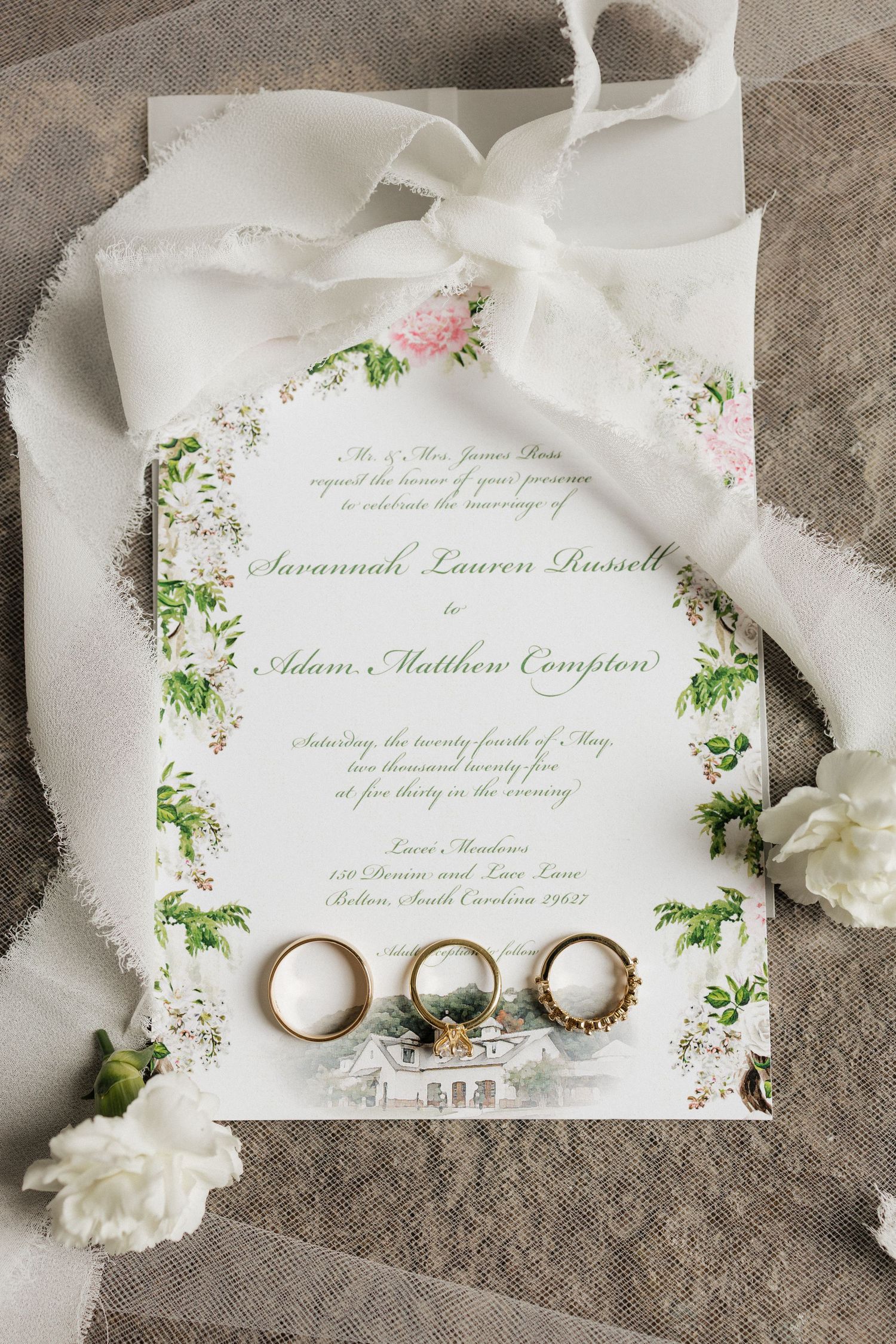 Wedding invitation with delicate floral border design alongside wedding rings and white fabric on gray stone surface.