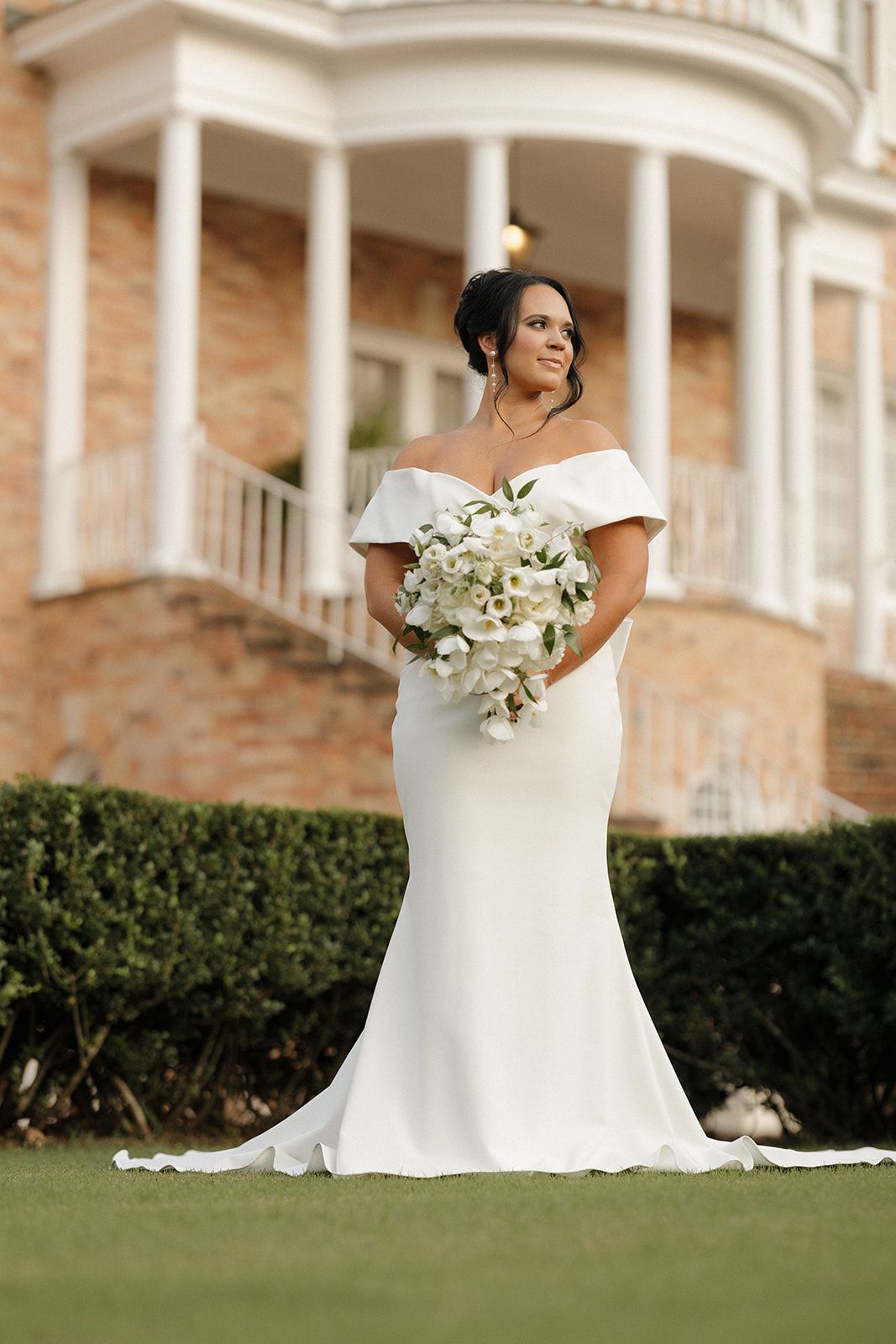 Elegant bride in off-shoulder mermaid wedding gown holding white bouquet poses on lawn in front of columned mansion.