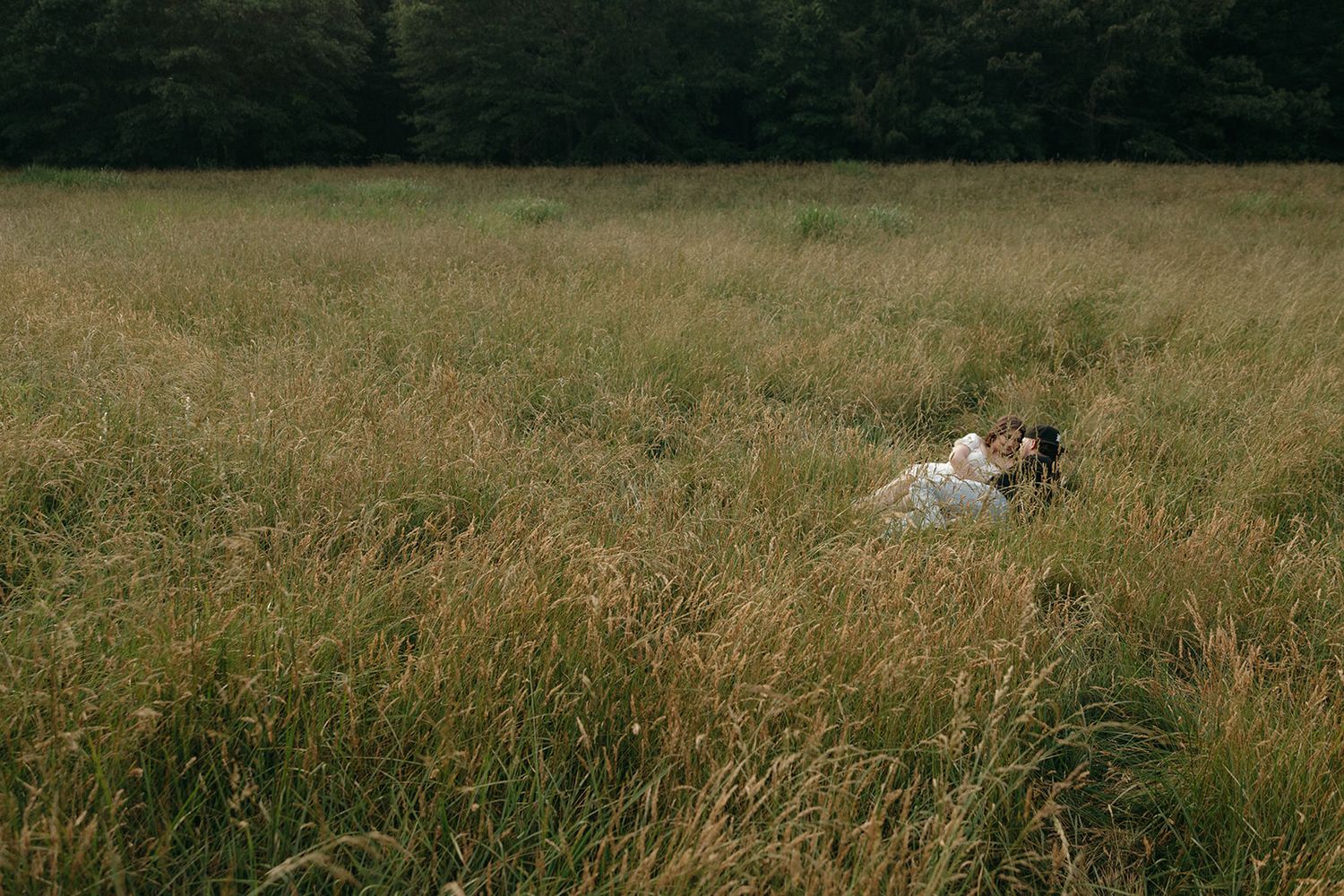 Two people relax in a peaceful meadow with tall grass and dark forest in the background.