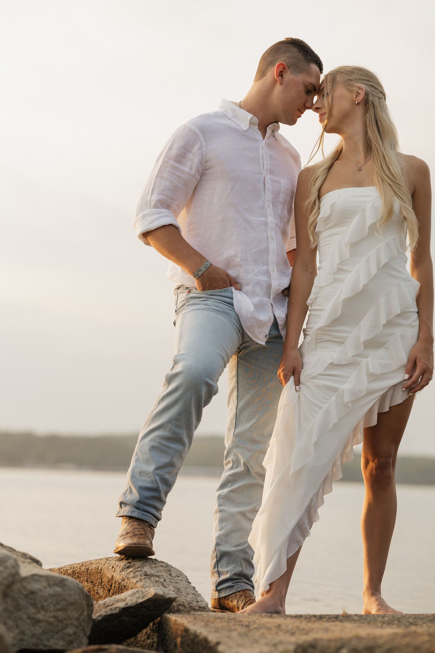 A romantic couple stands on rocky cliffs at sunset, photographed from multiple angles by the water.