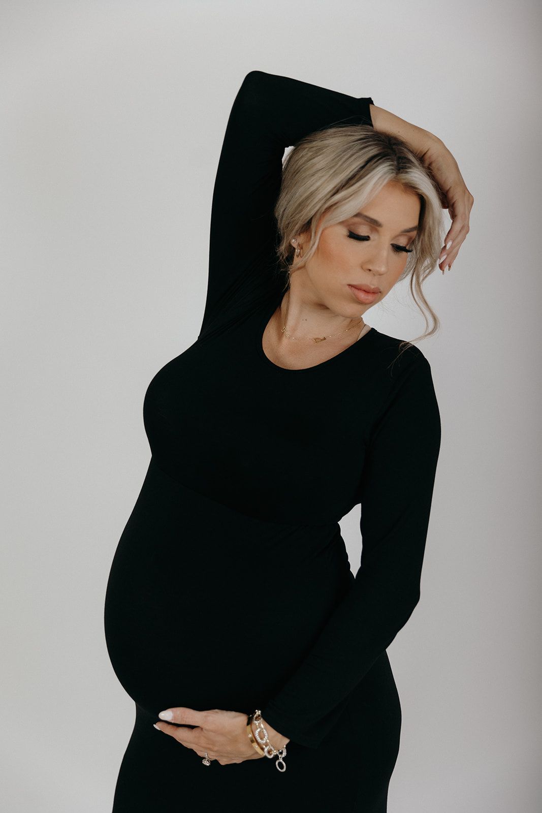 Elegant maternity photo of woman in black dress striking artistic pose against light gray background.