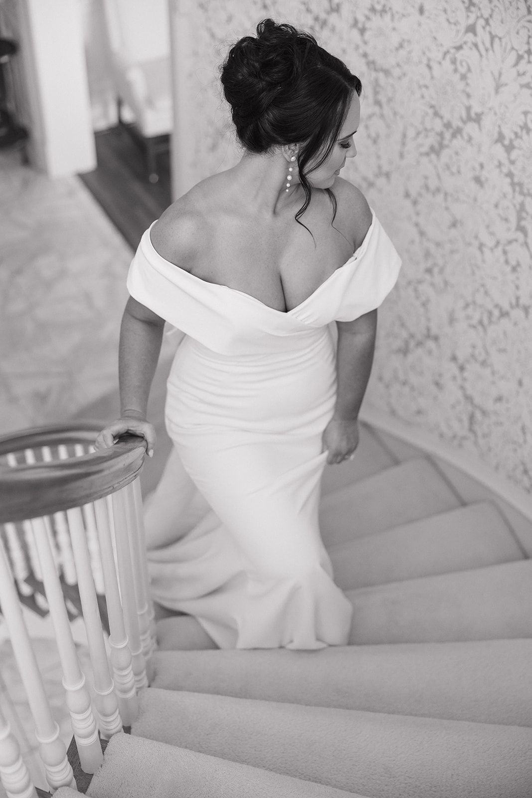 Elegant black and white photo of bride in off-shoulder wedding gown descending ornate staircase with vintage banister.