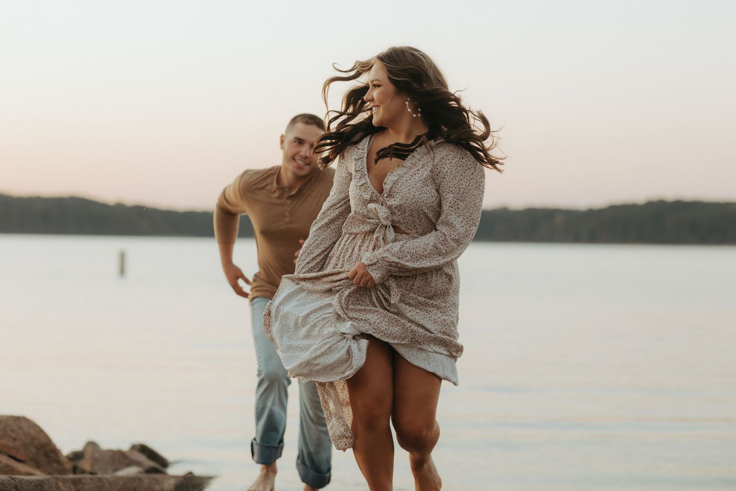 Playful couple running together by a serene lake at sunset with flowing dress and natural movement.