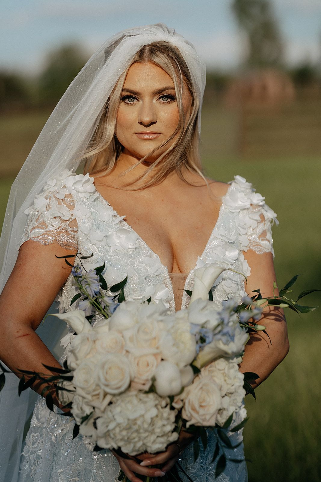 Beautiful bride in a lace wedding dress holds an elegant white and blue floral bouquet in an outdoor setting.