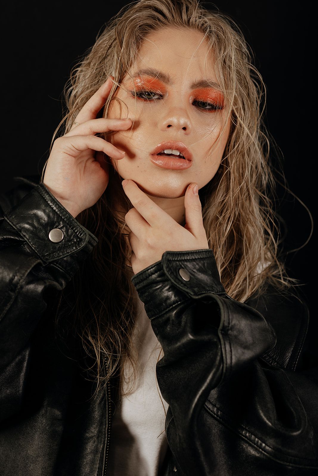 Fashion portrait in moody lighting with dramatic orange eye makeup and black leather jacket against dark background.
