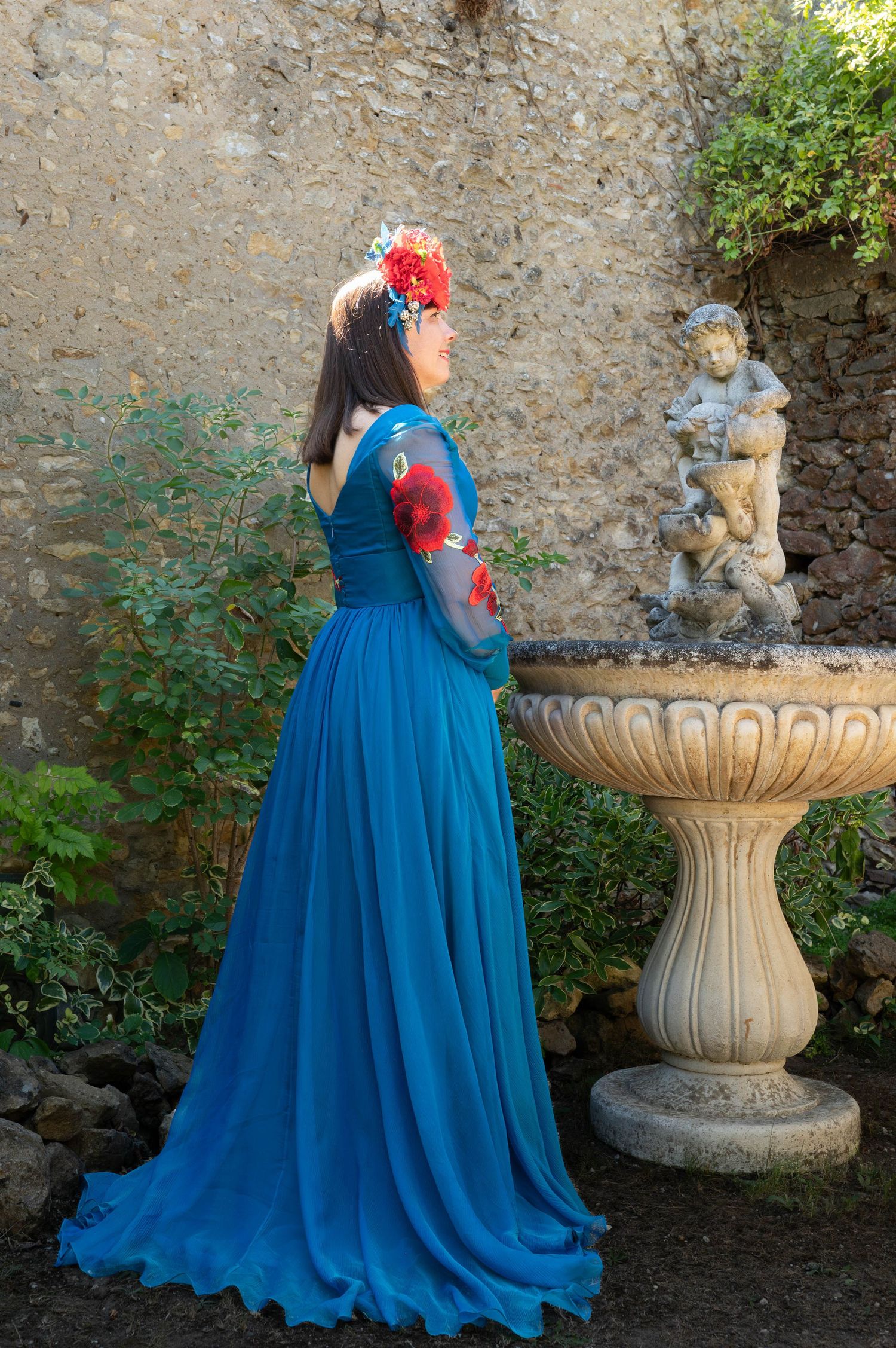 Long blue dress photographed against stone wall with ornate garden fountain.