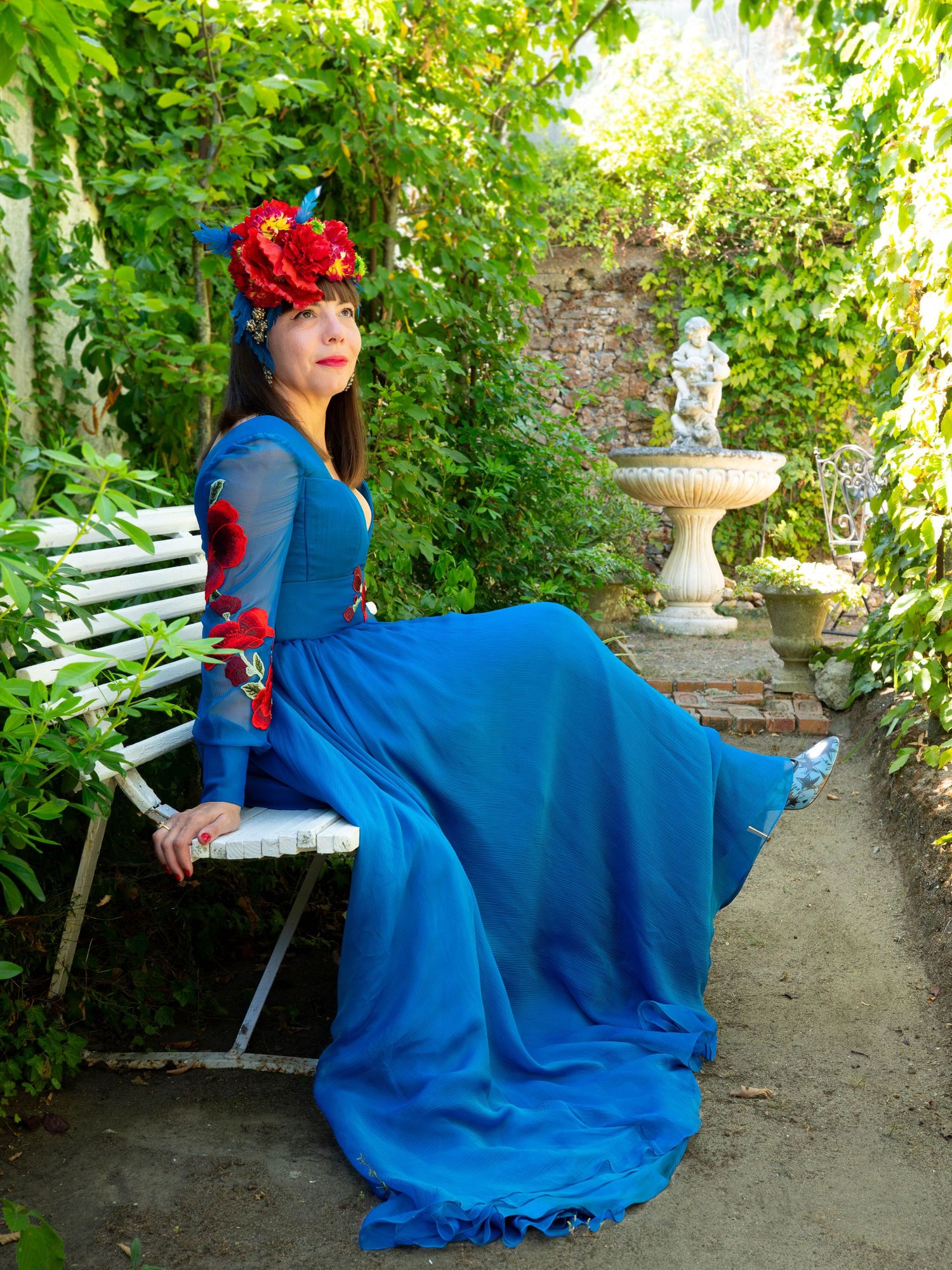 Woman in a vibrant blue gown sitting on a garden bench surrounded by lush greenery.