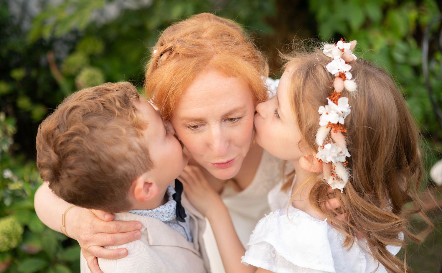 A tender family moment captured outdoors with a floral crown and natural greenery in the background.
